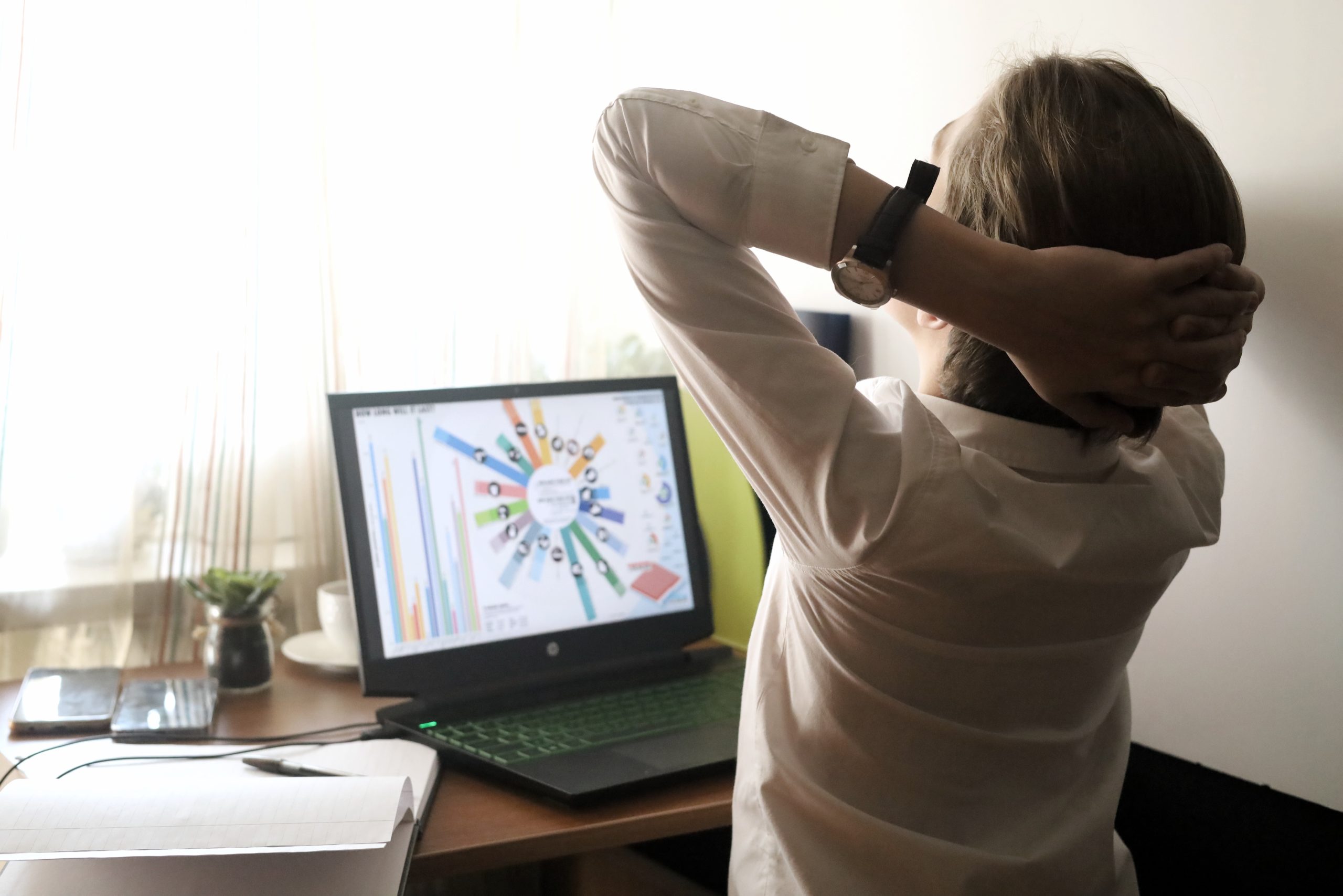A person in a white shirt stretches in front of a laptop displaying a colorful graph, amidst notebooks and a phone, embodying the art of managing screen time for better wellbeing.