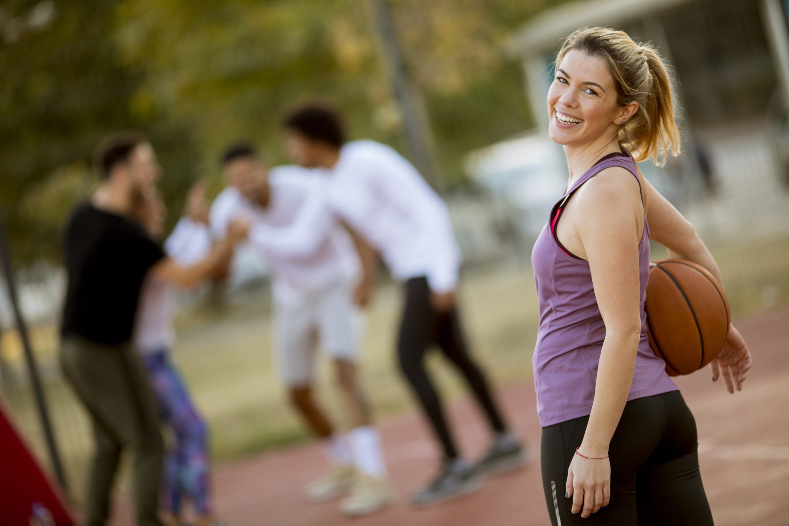 A woman holding a basketball stands on an outdoor court, smiling at the camera, radiating a positive mood, while a group of people interact in the background.