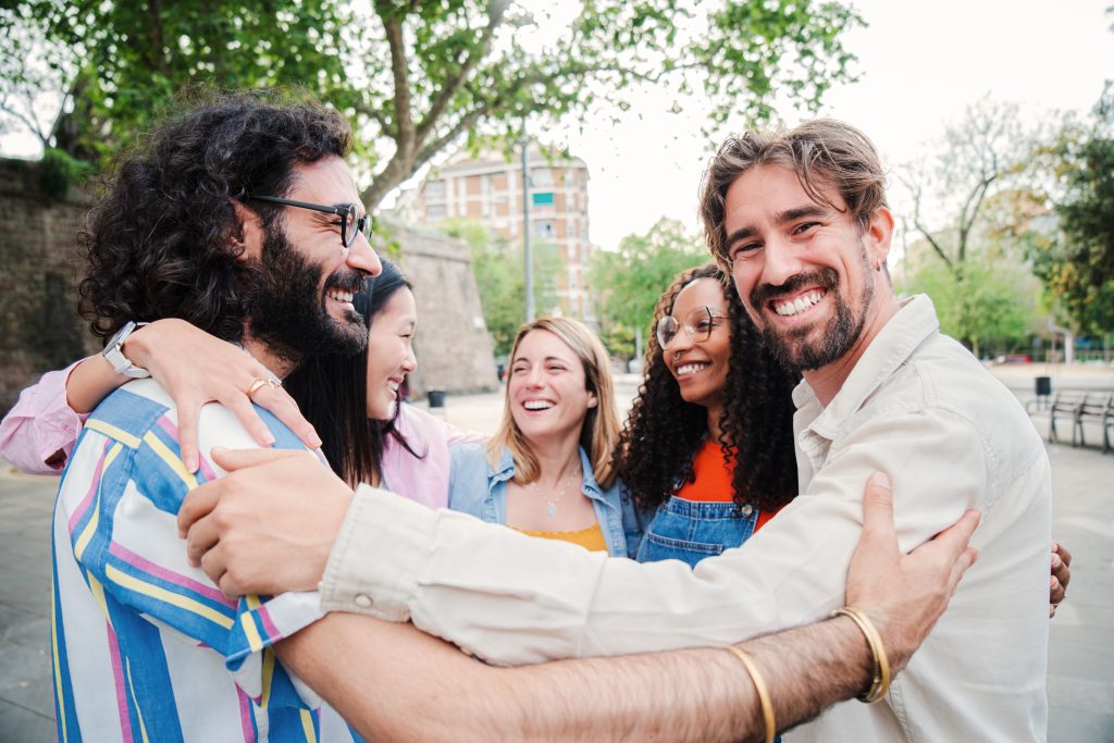Five people stand outdoors, smiling and hugging each other in a park setting with trees and buildings in the background, capturing the perfect post-worthy moment of joy together.