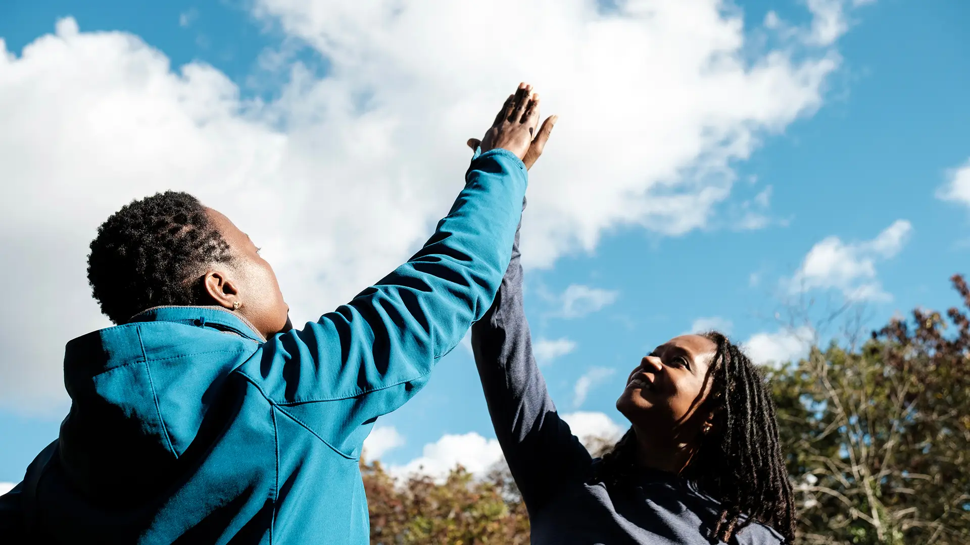 Two people outdoors giving a high-five under a cloudy blue sky. They are both wearing jackets.