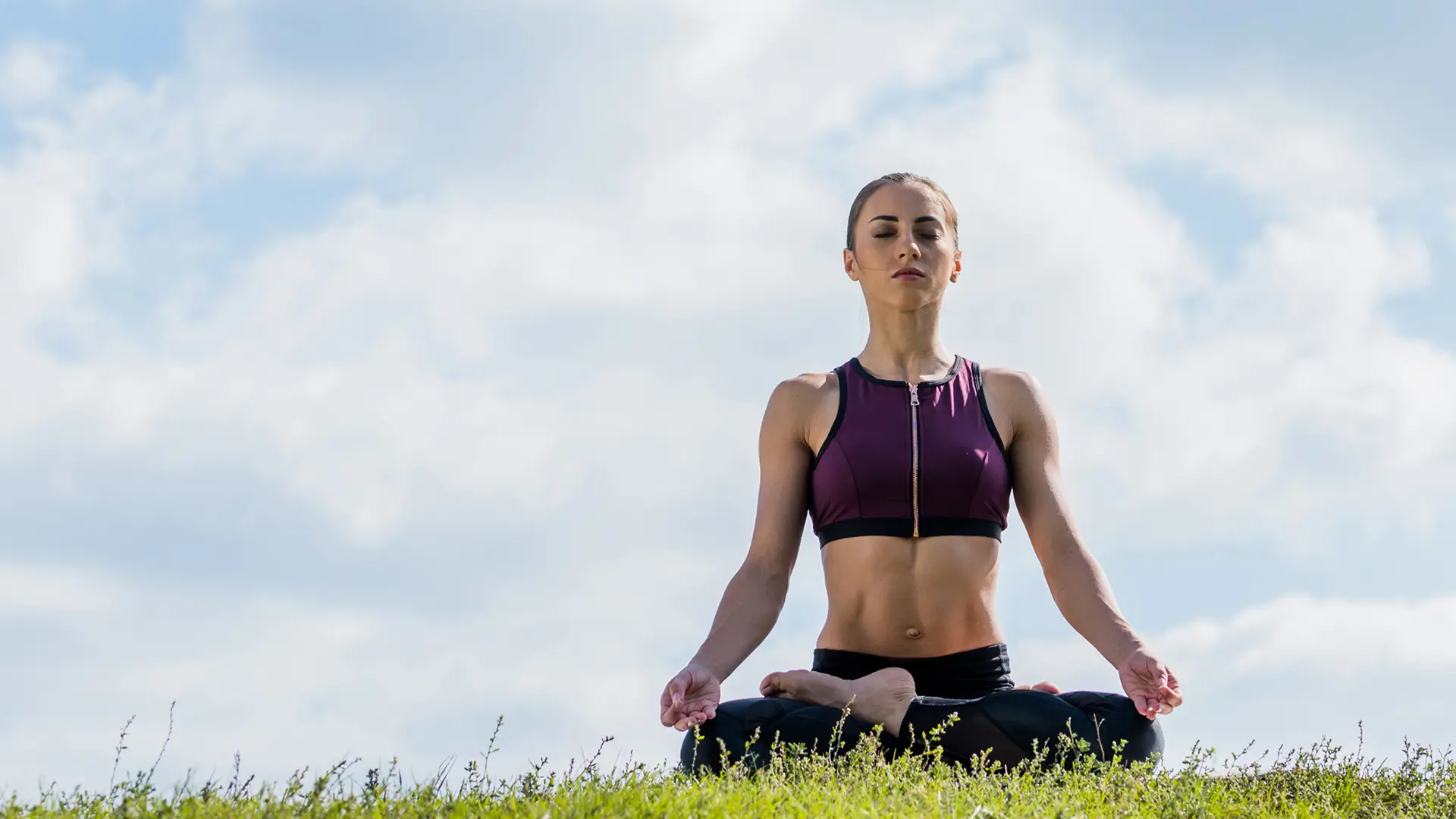 A person sits cross-legged on grass, meditating under a cloudy sky, wearing athletic clothing.
