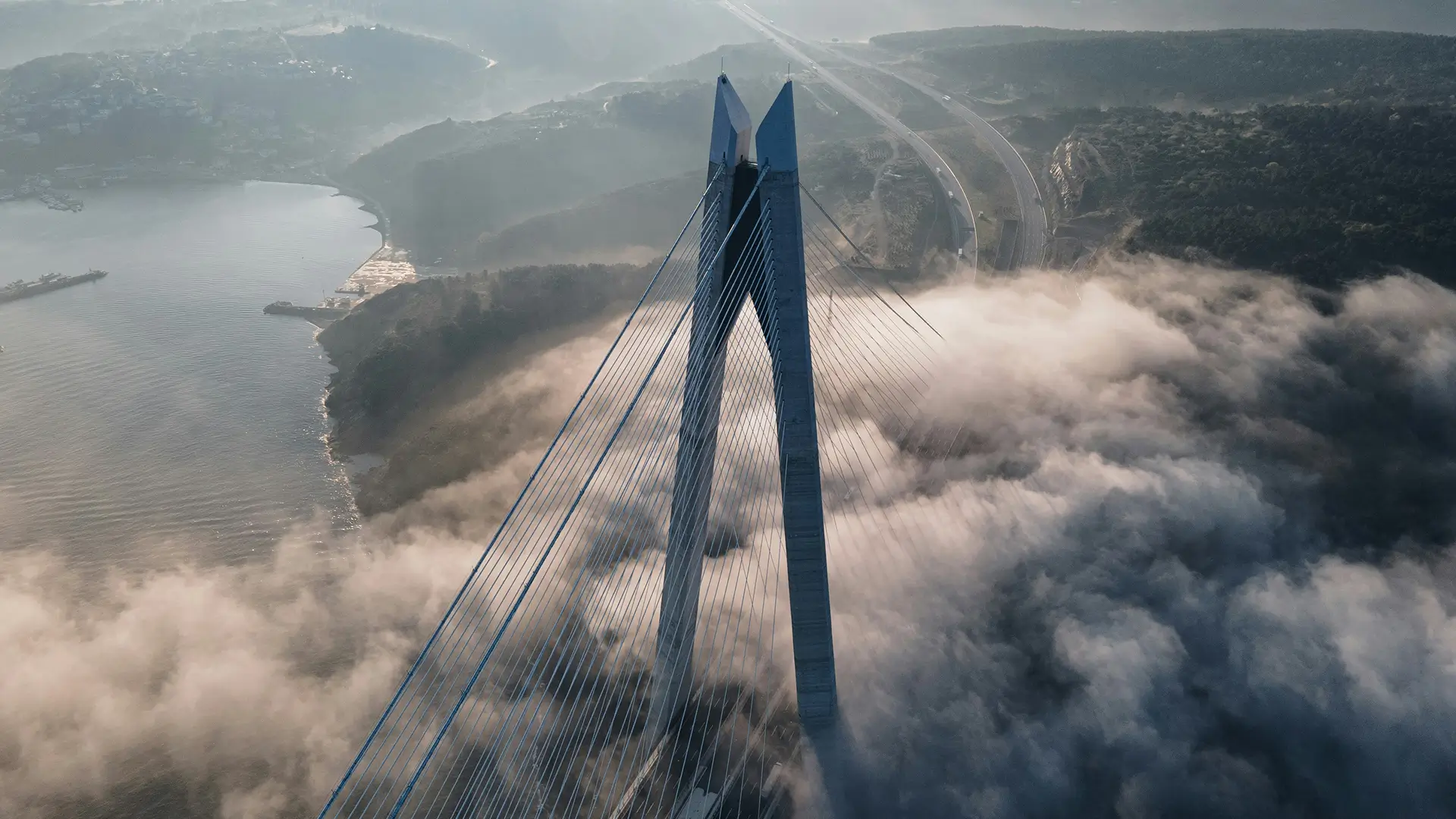A suspension bridge rises above fog-covered water and land, with visible cables and distant roads and hills under a cloudy sky.