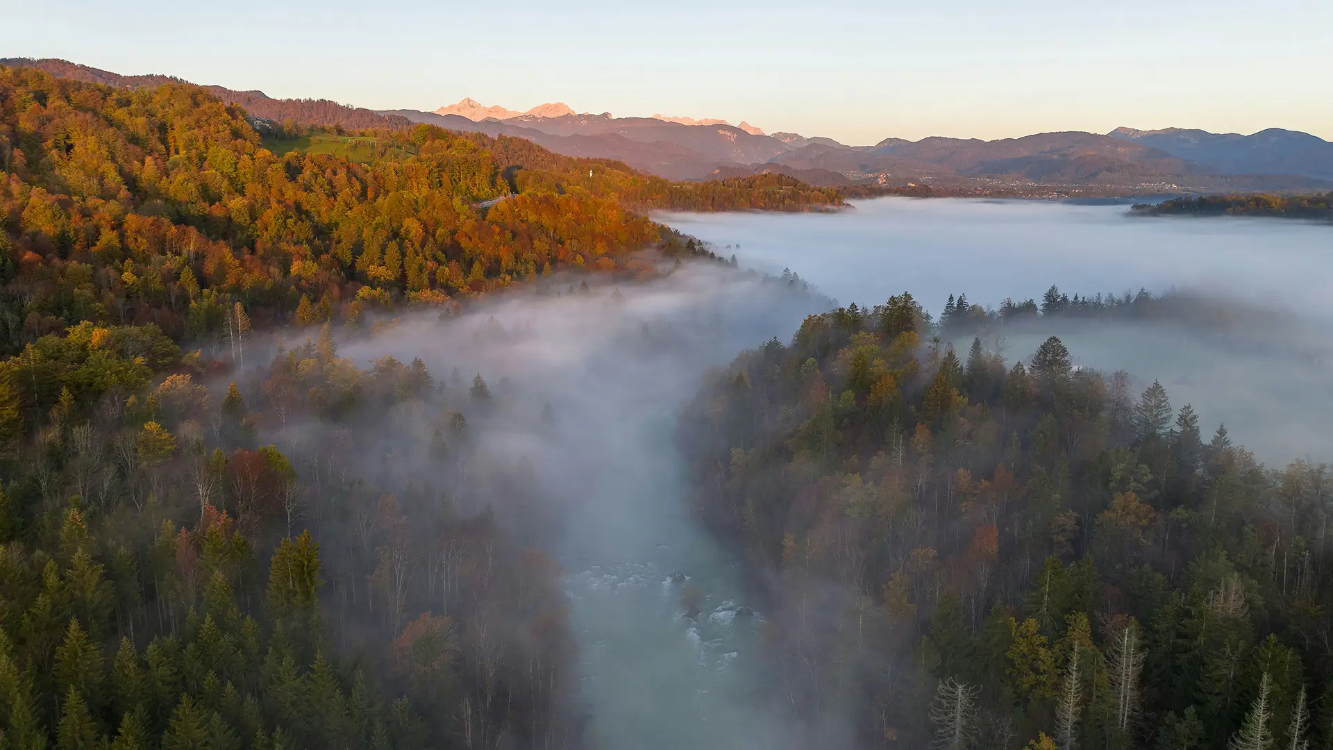 Aerial view of a mist-covered river cutting through a forested valley, surrounded by rolling hills. Trees display autumn colors, and distant mountains are visible on the horizon.
