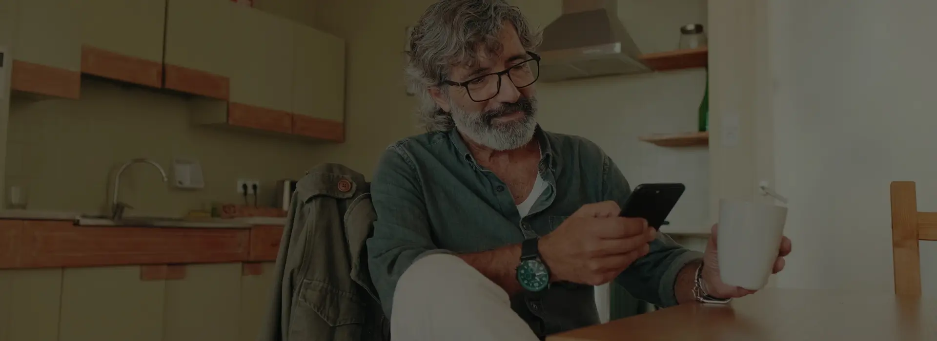 Man with gray hair and glasses sitting at a table using a smartphone, holding a white mug, with a kitchen in the background.
