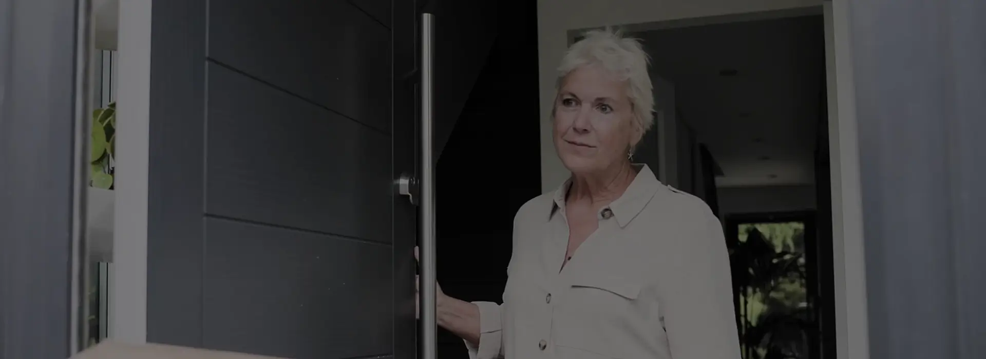 A woman with short white hair stands at a partially open door in a modern interior setting, wearing a light-colored blouse.