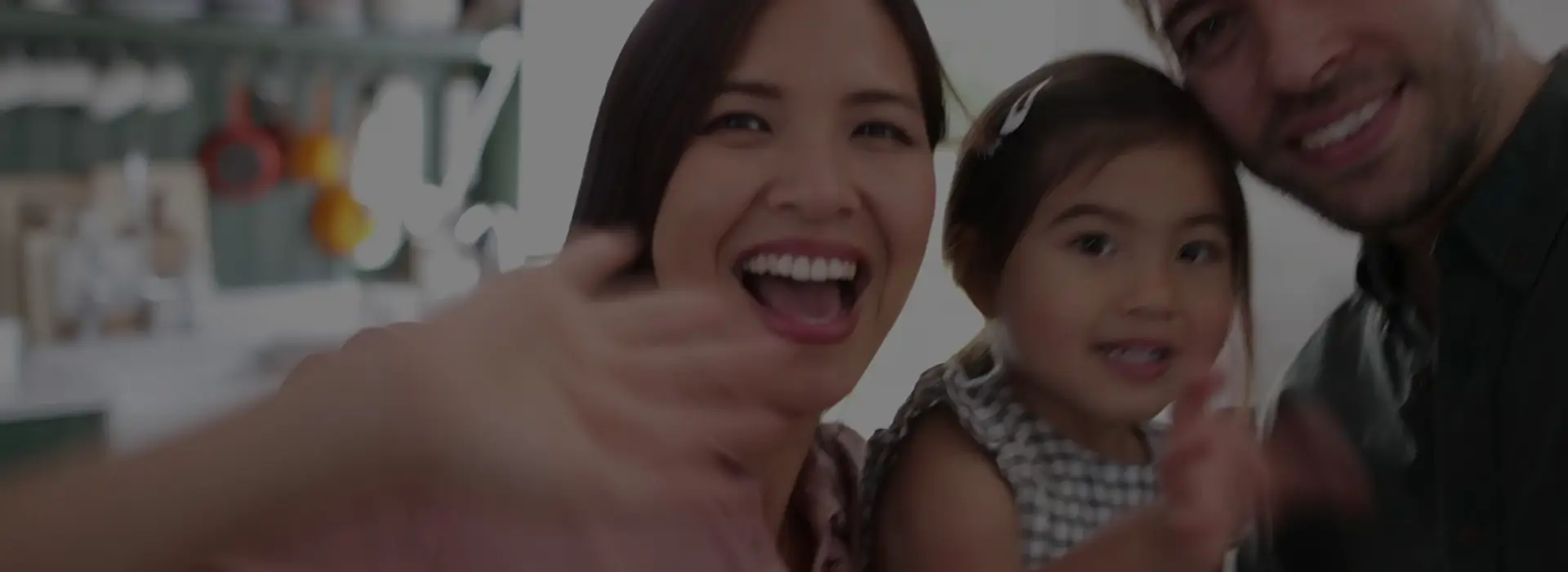 A family of three smiling and waving at the camera, with the mother holding the child in the foreground.
