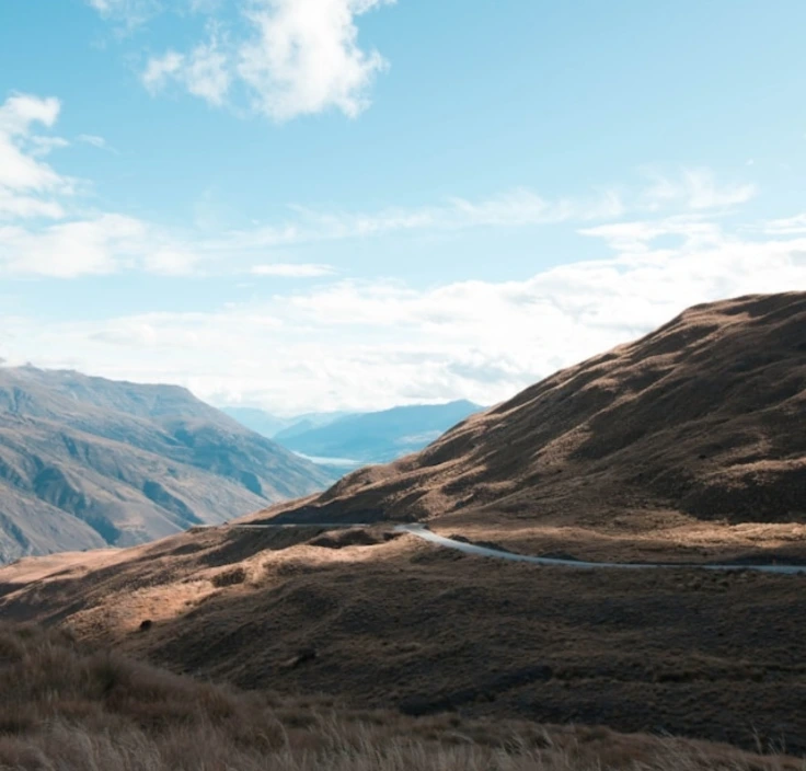 A winding road cuts through dry, rolling hills under a clear blue sky with distant mountains in the background.