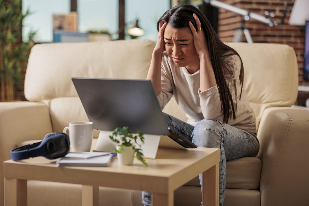 A woman sits on a couch, gripping her head in frustration as she stares at her laptop. Surrounded by a plant, cup, headphones, and notebook on the table, she's clearly in need of a digital detox to reclaim her sense of wellbeing.