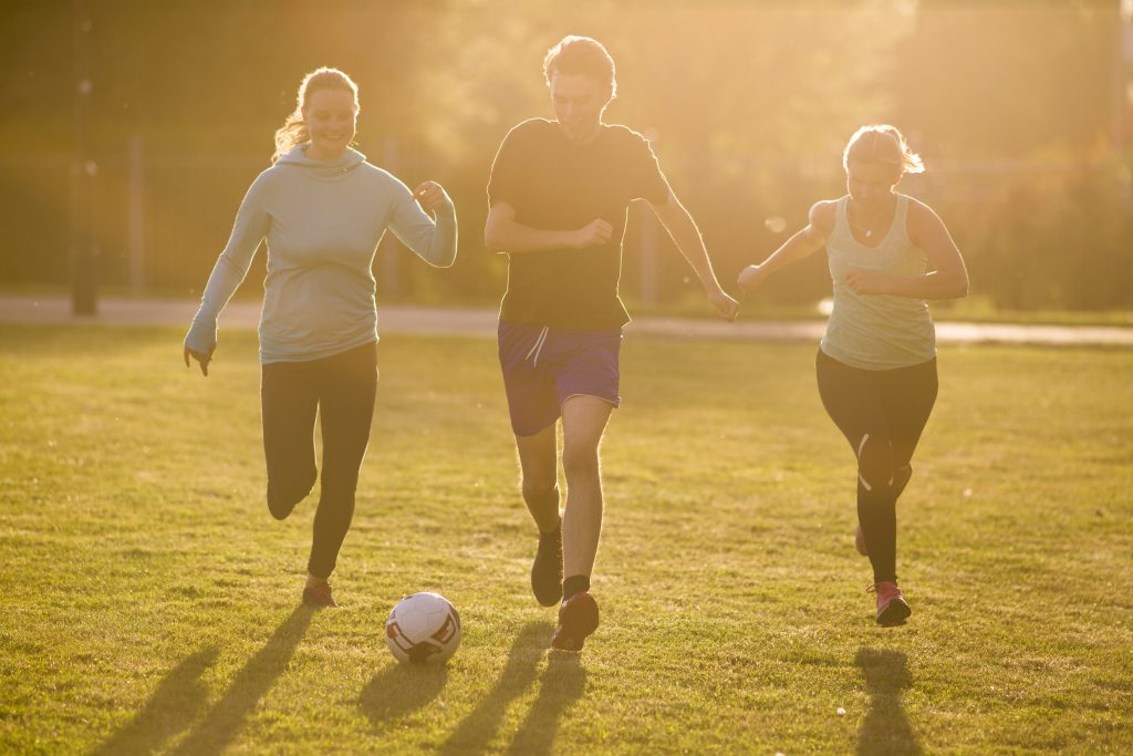 Three people enjoy a lively soccer game on a grassy field under the sunlight, with one person skillfully kicking the ball while maintaining balance and the others running nearby, fully immersed in the invigorating physical activity.